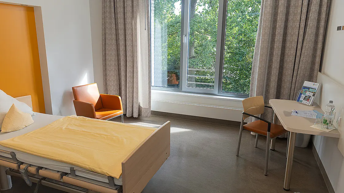 Ein Reha-Zimmer aus dem Regelleistungsbereich mit einem Bett, einem Sessel und einem Schreibtisch.