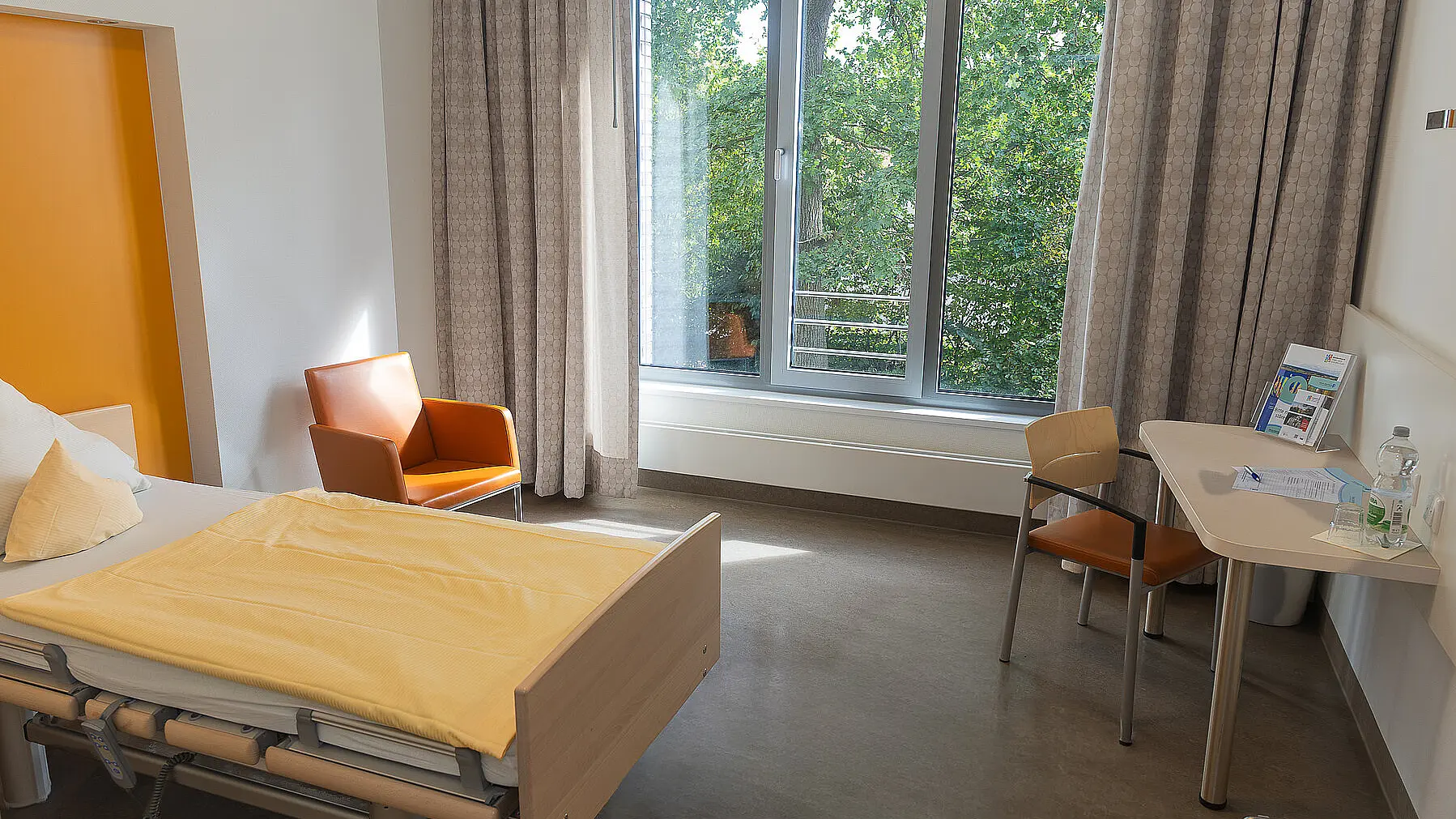 Ein Reha-Zimmer aus dem Regelleistungsbereich mit einem Bett, einem Sessel und einem Schreibtisch.