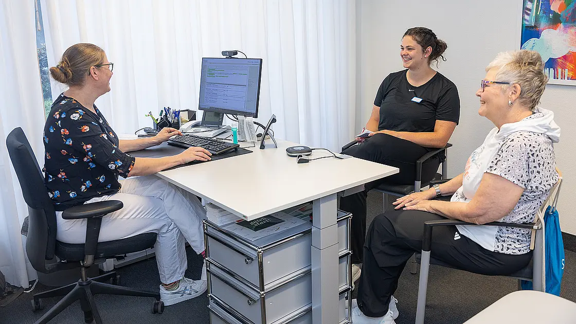 Eine Ärztin und eine Physiotherapeutin sitzen mit einer Patientin im Besprechungszimmer.
