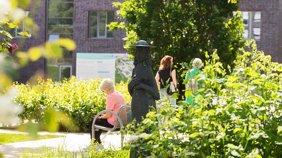 Eine Frau sitzt auf einer Bank und liest, zwei andere Menschen gehen vorbei und spazieren gemeinsam durch den Park. Im Hintergrund ist das Reha-Zentrum zu sehen.