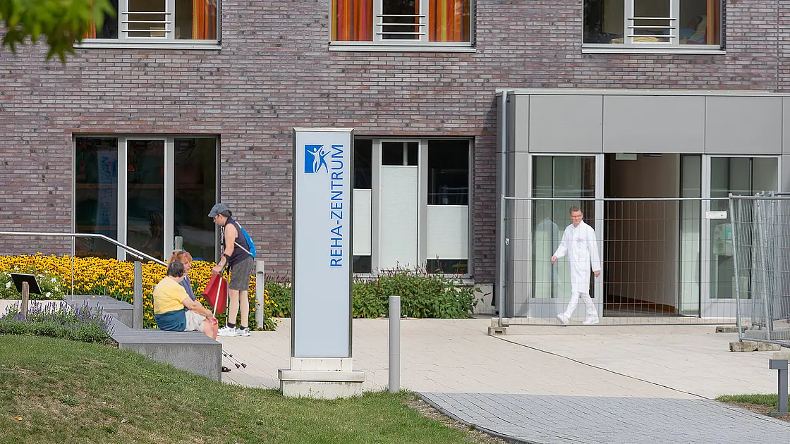 Ein Foto vonn dem Reha-Schild, im Hintergrund sitzen Menschen mit Gehhilfen auf einer Treppe und ein Arzt läuft aus dem Eingang heraus.