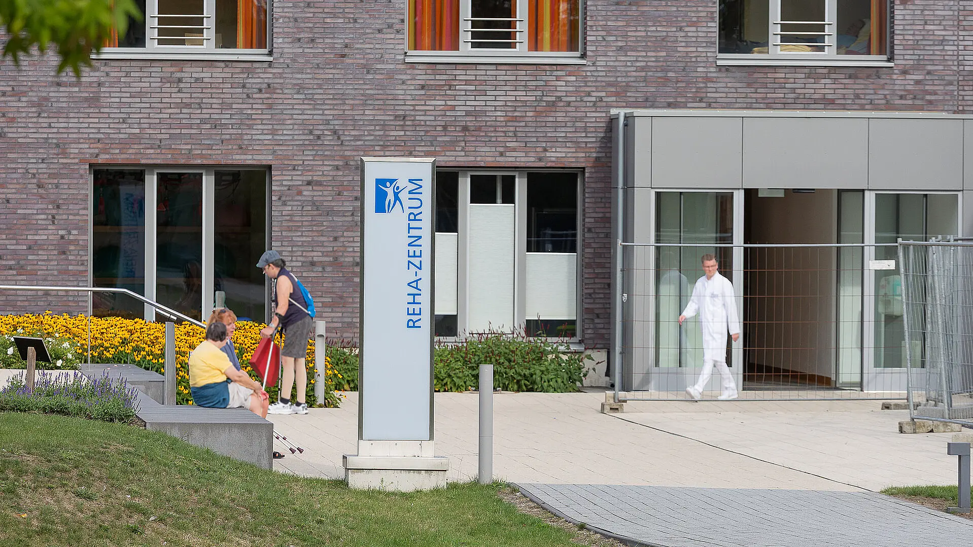 Ein Foto vonn dem Reha-Schild, im Hintergrund sitzen Menschen mit Gehhilfen auf einer Treppe und ein Arzt läuft aus dem Eingang heraus.