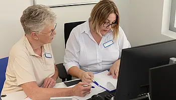 Zwei Mitarbeiterinnen besprechen Unterlagen an einem Arbeitsplatz mit Computer.