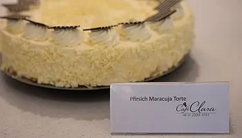 Eine Pfirsich-Maracuja-Torte mit Sahnetupfen und Schokoladendekor steht auf einer Servierplatte. Davor befindet sich ein Schild mit der Aufschrift „Pfirsich Maracuja Torte“.
