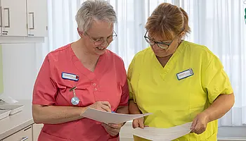 Zwei Pflegekräfte stehen nebeneinander und besprechen gemeinsam Unterlagen.