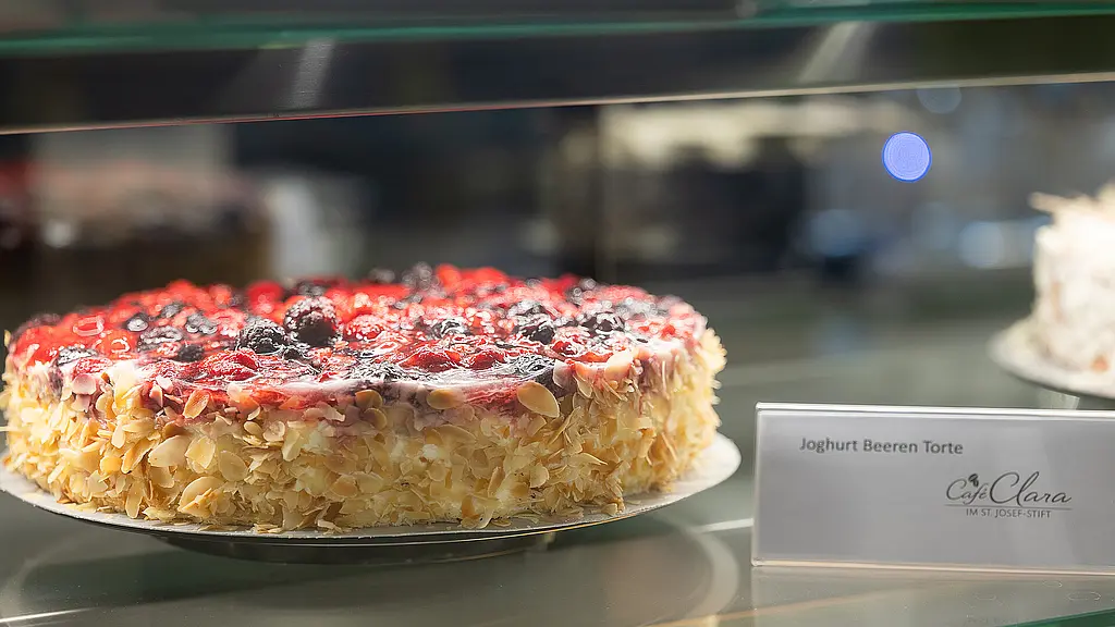Eine Joghurt-Beeren-Torte mit Mandelrand und frischen Beeren steht in einer Kuchenvitrine. Daneben befindet sich ein Schild mit der Produktbezeichnung.