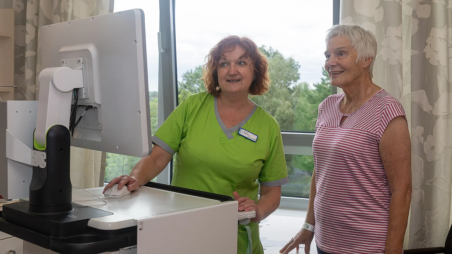 Eine Pflegekraft und eine Patientin stehen gemeinsam vor einem Computerbildschirm im Patientenzimmer.