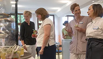 Mehrere Gäste und Mitarbeitende stehen an der Verkaufstheke des Café Clara, betrachten das Angebot und kommen miteinander ins Gespräch.