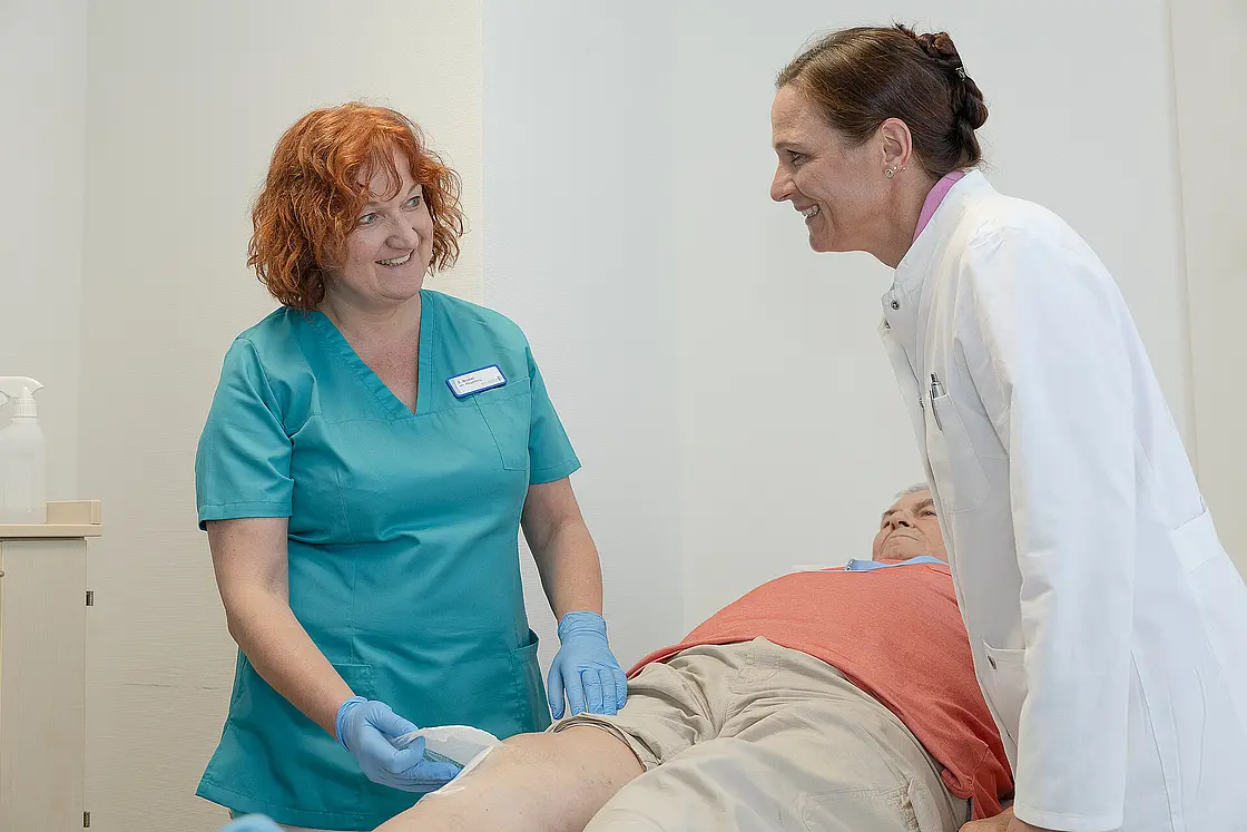 Eine Pflegekraft versorgt das Knie eines liegenden Patienten, während eine Ärztin unterstützt.