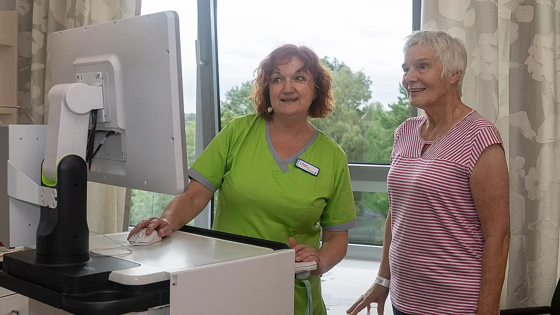 Eine Pflegekraft und eine Patientin stehen gemeinsam vor einem Computerbildschirm im Patientenzimmer.
