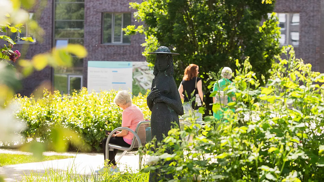 Eine Frau sitzt auf einer Bank und liest, zwei andere Menschen gehen vorbei und spazieren gemeinsam durch den Park. Im Hintergrund ist das Reha-Zentrum zu sehen.