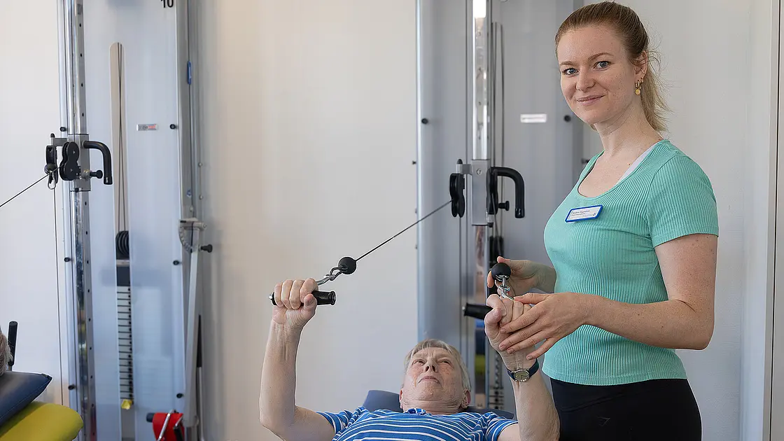Eine Patientin trainiert im Liegen an einem Seilzuggerät, während eine Physiotherapeutin die Übung begleitet.