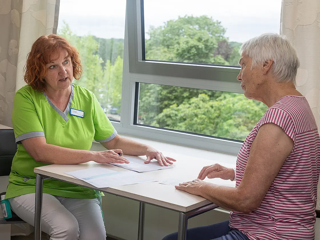 Eine Pflegekraft sitzt einer Patientin am Tisch gegenüber und macht sich während des Gesprächs Notizen.