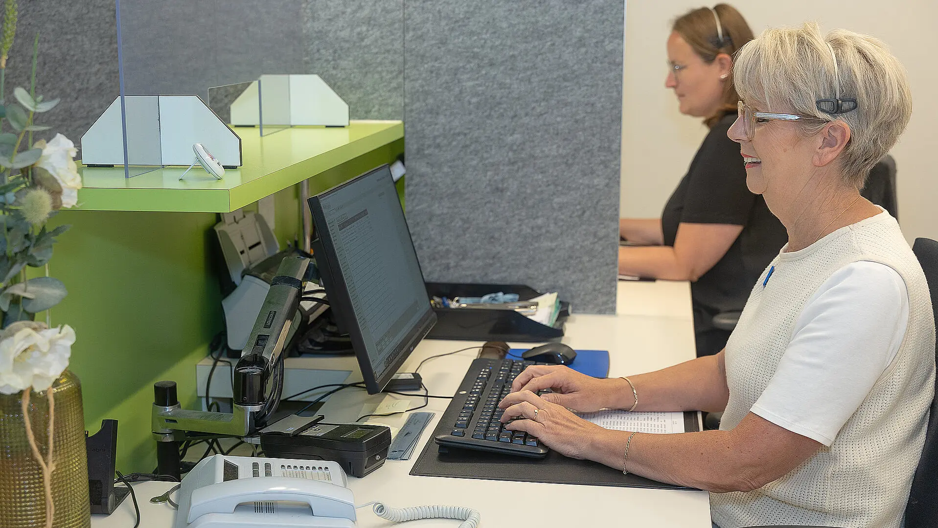 Zwei Mitarbeiterinnen des Reha-Patientenmanagements bearbeiten Anträge von extern vor einem PC im Empfangsbereich der Reha.