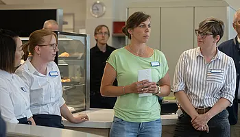 Eine Mitarbeiterin spricht vor einer kleinen Gruppe von Gästen und Mitarbeitenden im Verkaufsbereich des Café Clara.