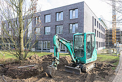 Bagger arbeitet im Außenbereich vor dem Neubau des Reha-Zentrums; daneben ein mehrgeschossiges Gebäude mit Backsteinfassade und eingerüsteten Bereichen im Hintergrund
