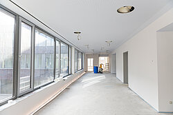 Heller, langer Flur im Neubau der Reha mit großen Fensterfronten links, noch unfertiger Decke mit sichtbaren Kabelanschlüssen und leerem Boden; Baumaterialien stehen im Hintergrund